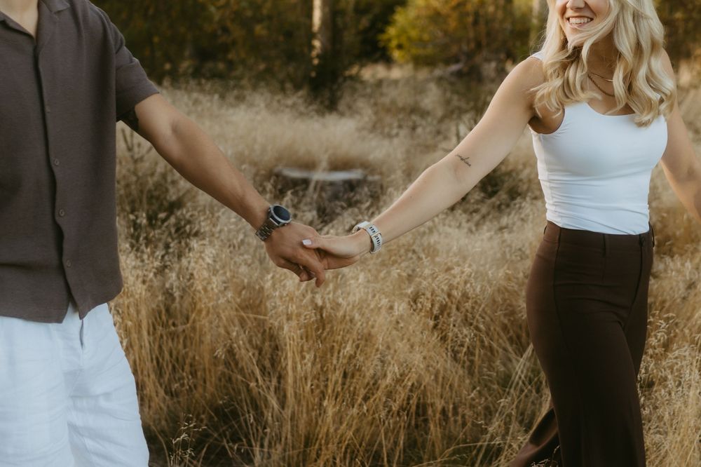 Couple holding hands while walking through a golden grassy field, the woman in a white tank top smiling in warm natural light.