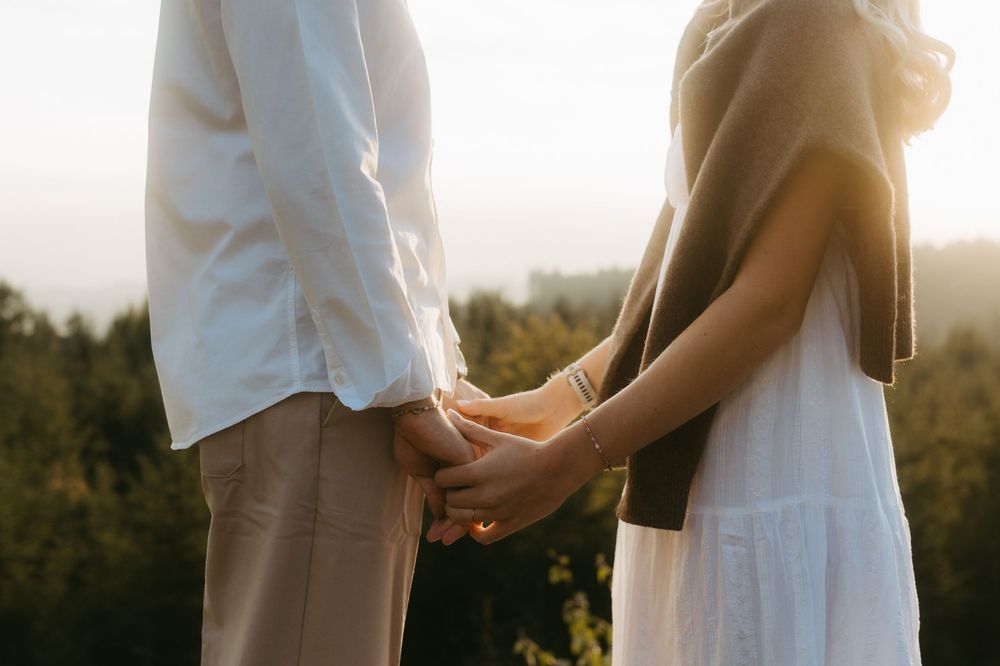 Couple holding hands outdoors at sunset, standing in front of a forested hillside with warm golden light.