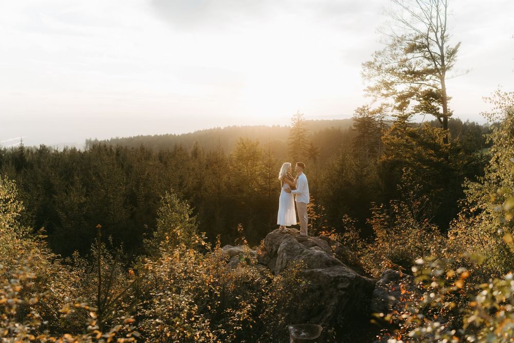 Couple in white embracing on a rocky outcrop overlooking a golden-lit pine forest at sunset.