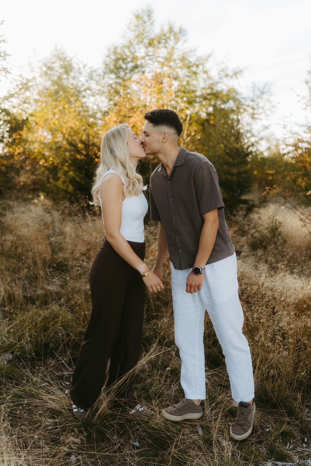 Couple sharing a kiss while holding hands in a golden grassy field with autumn trees, a romantic outdoor engagement photo.