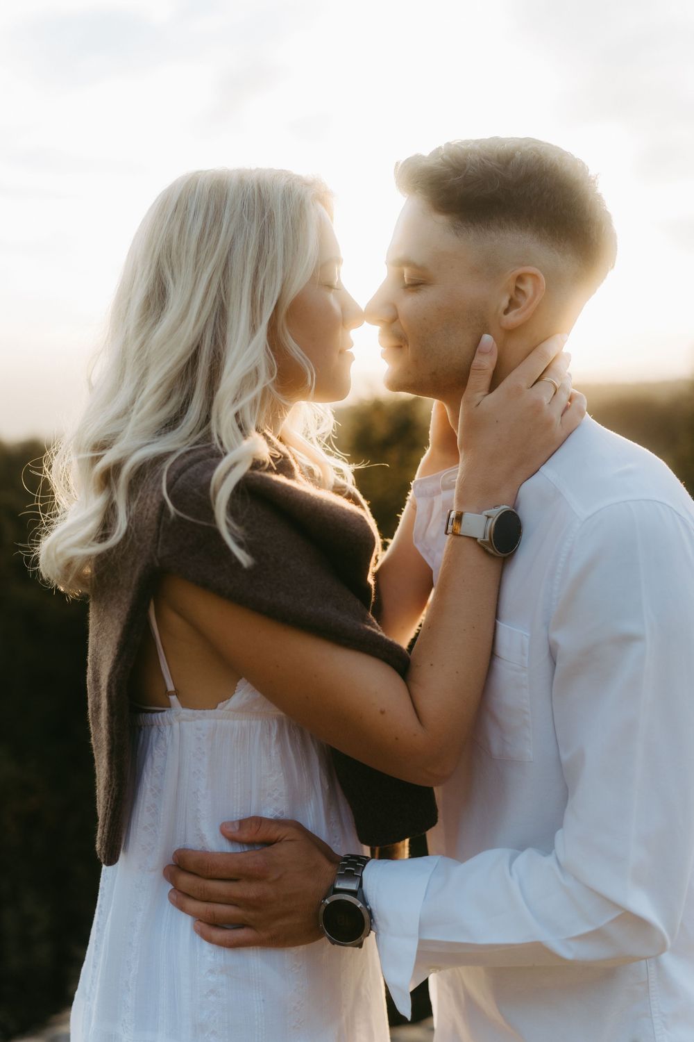 Romantic couple in white outfits nearly kiss at sunset, the man cupping the woman's neck as she wears a brown sweater.