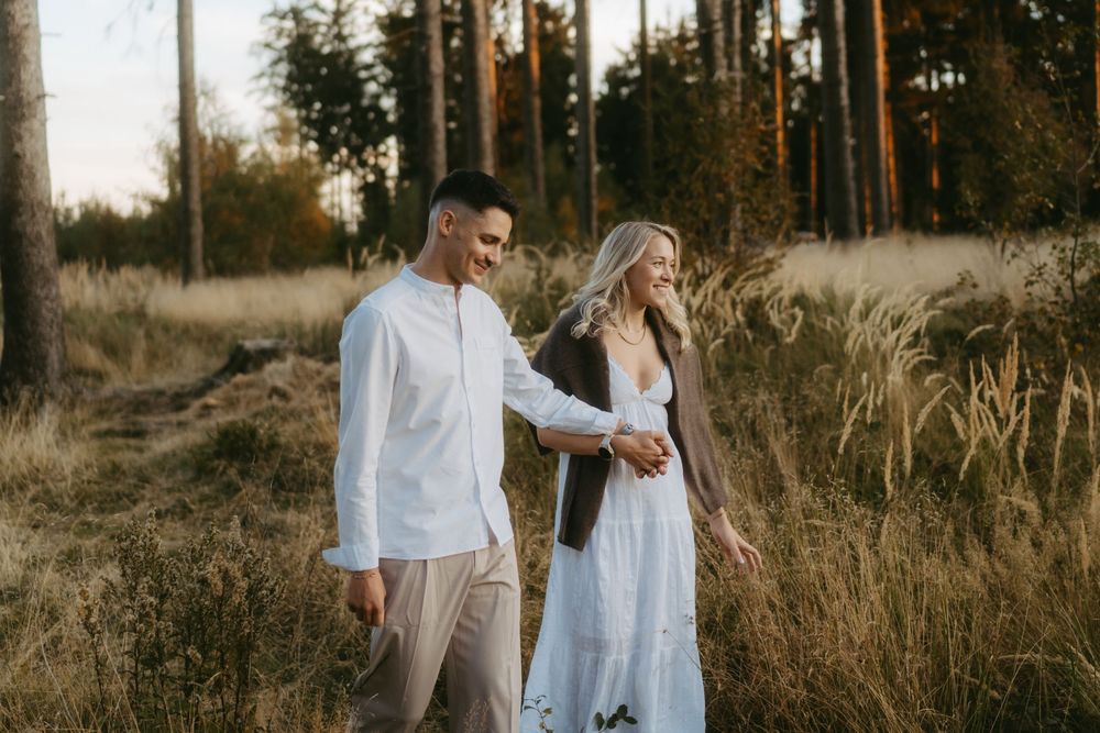 Couple in white outfits holding hands and laughing while walking through tall grass near a forest at sunset.