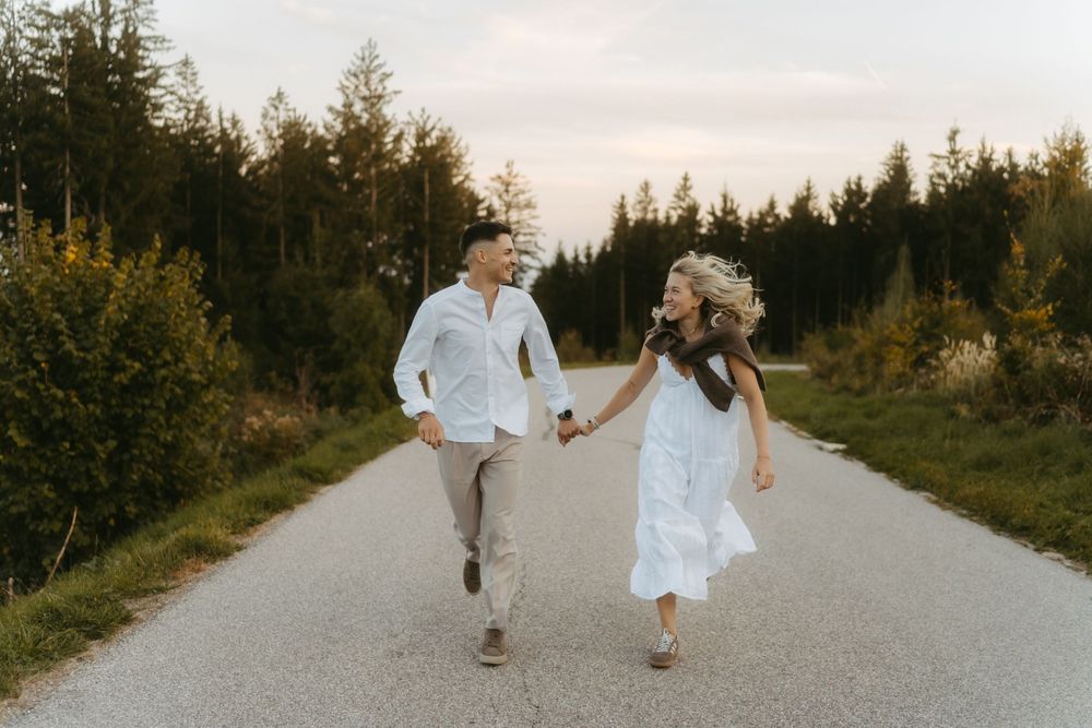 Smiling couple holding hands and running down a paved country road surrounded by green trees and grass at sunset.