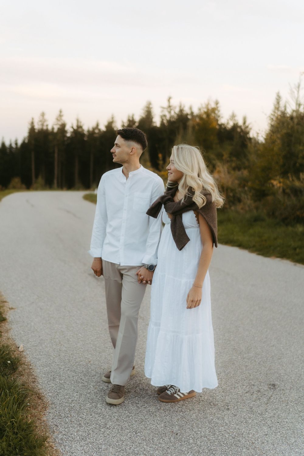 Couple holding hands on a quiet paved path lined with evergreen trees at dusk, the woman in a white dress.