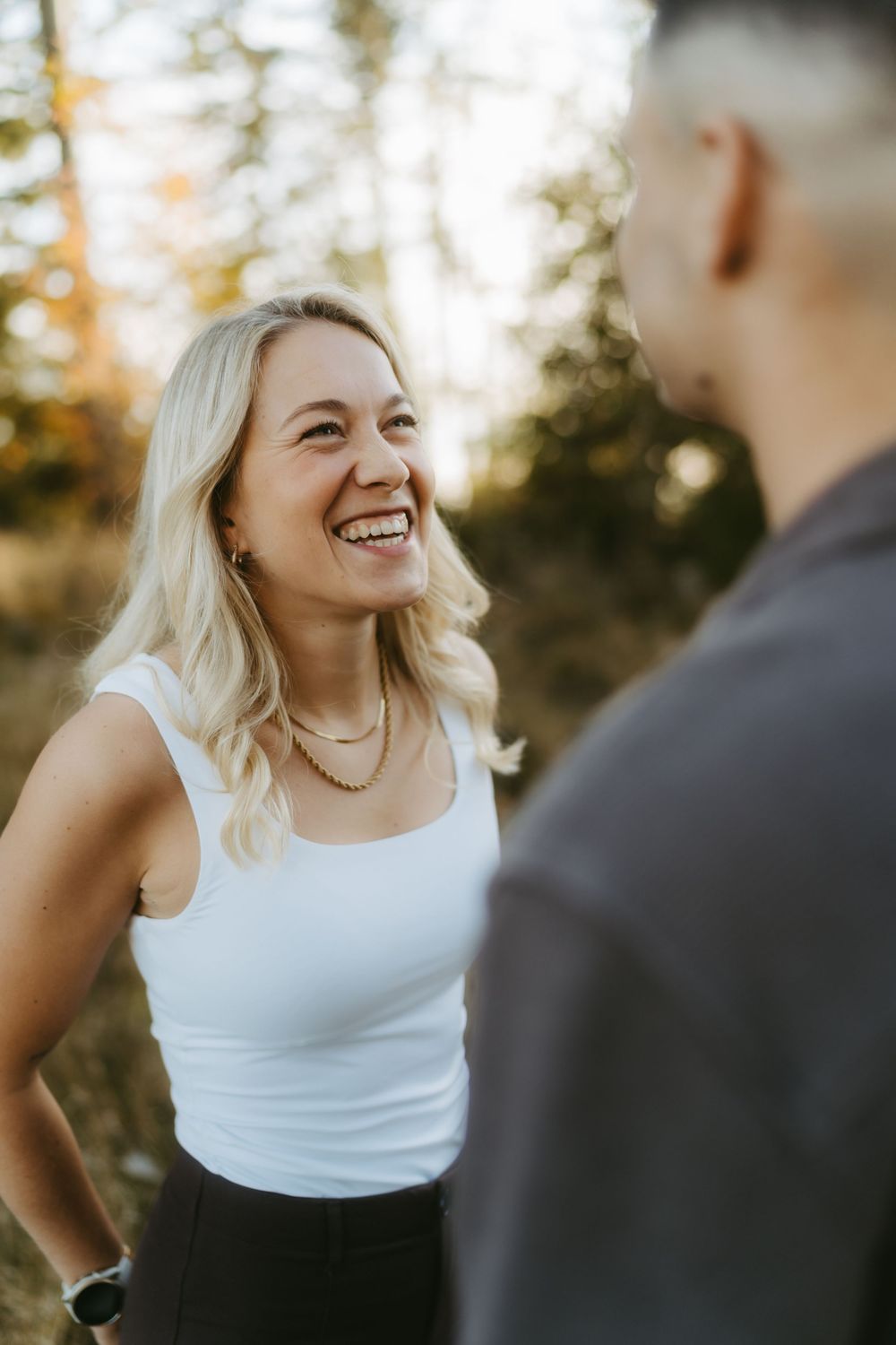Smiling blonde woman in a white tank top and layered necklaces laughs with a man in the foreground, outdoors among golden autumn trees.