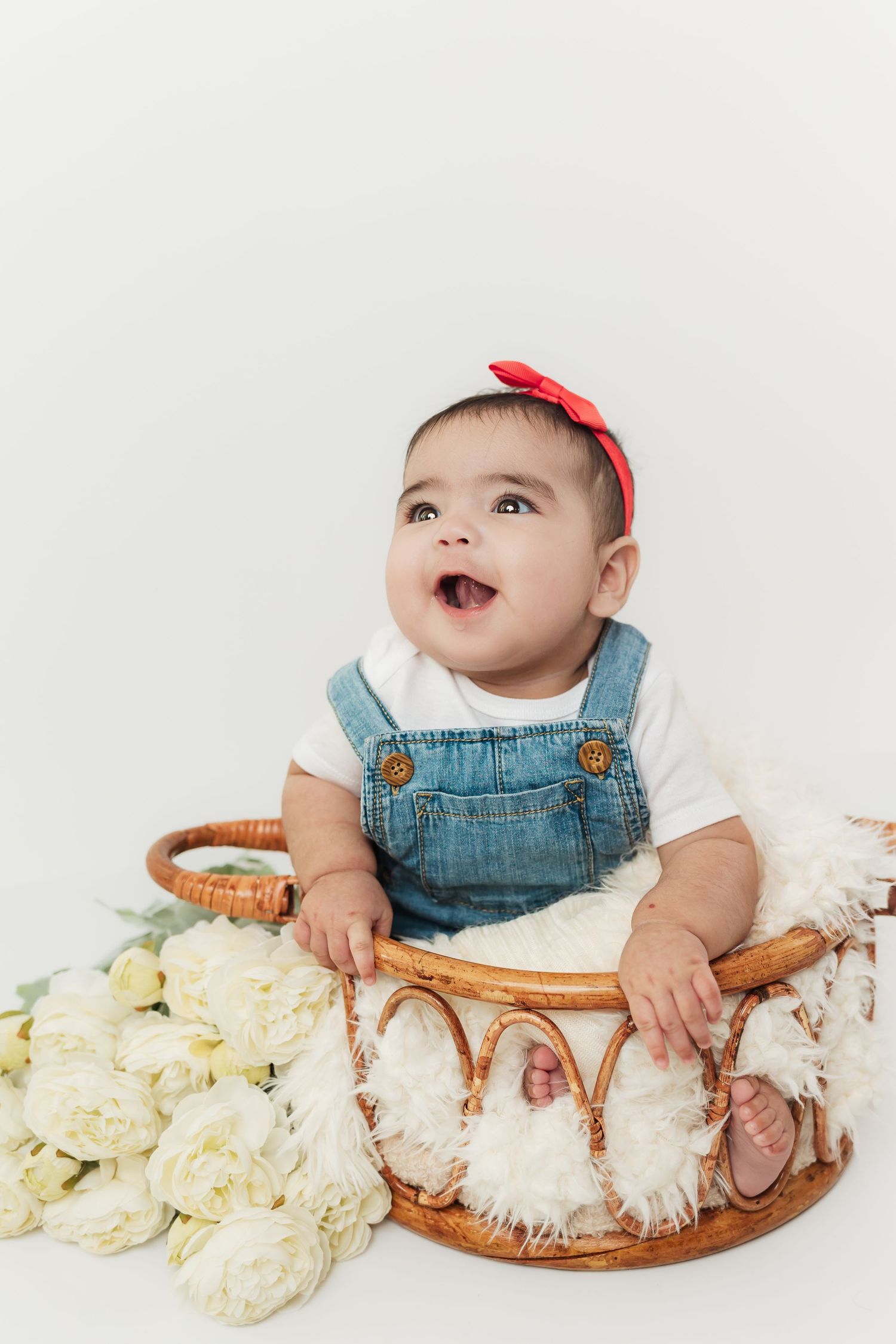 Adorable baby girl in denim overalls and red bow headband sitting in a wicker basket with white roses.