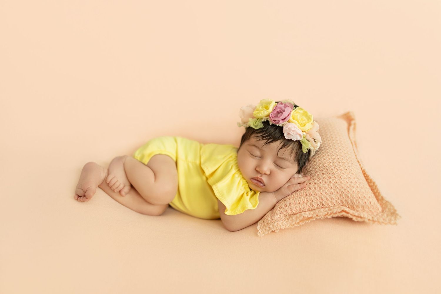 Sleeping newborn baby in yellow romper with floral headband resting on peach pillow against soft beige background.
