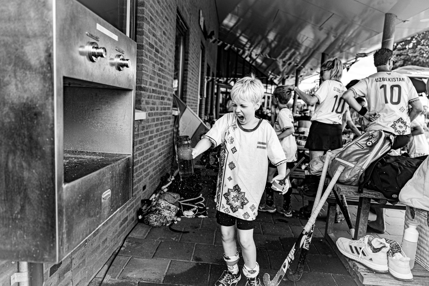 Black and white photo of a young blond child in sports jersey holding a flag near brick building with people in background.
