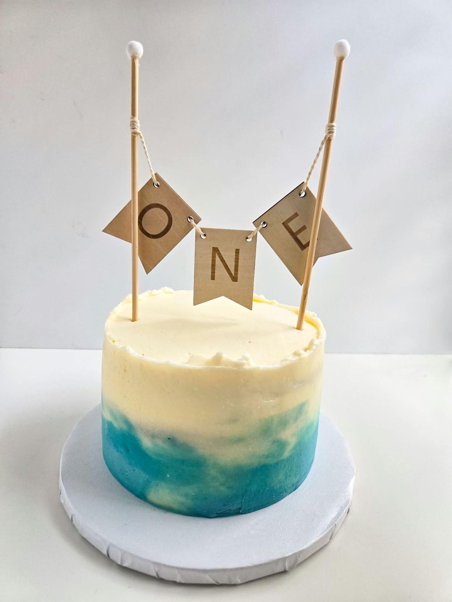 First birthday smash cake with blue watercolor frosting and wooden ONE banner topper on white cake board.