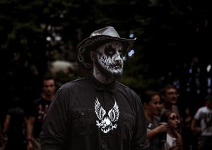 Person wearing skull face paint and cowboy hat at outdoor event, dressed in black graphic tee.