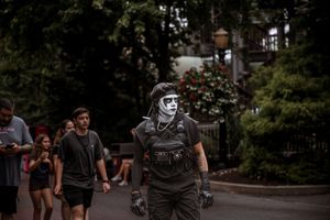 Person in dramatic face paint and tactical gear walks among crowd on tree-lined path at outdoor event.
