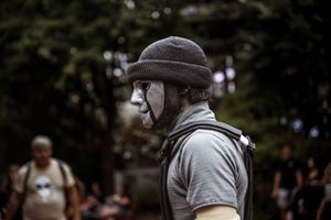 Man wearing a gray beanie hat and white theatrical mime mask outdoors among trees.