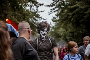 Person with dramatic black and white zombie face paint walks among crowd outdoors near tall evergreen trees.