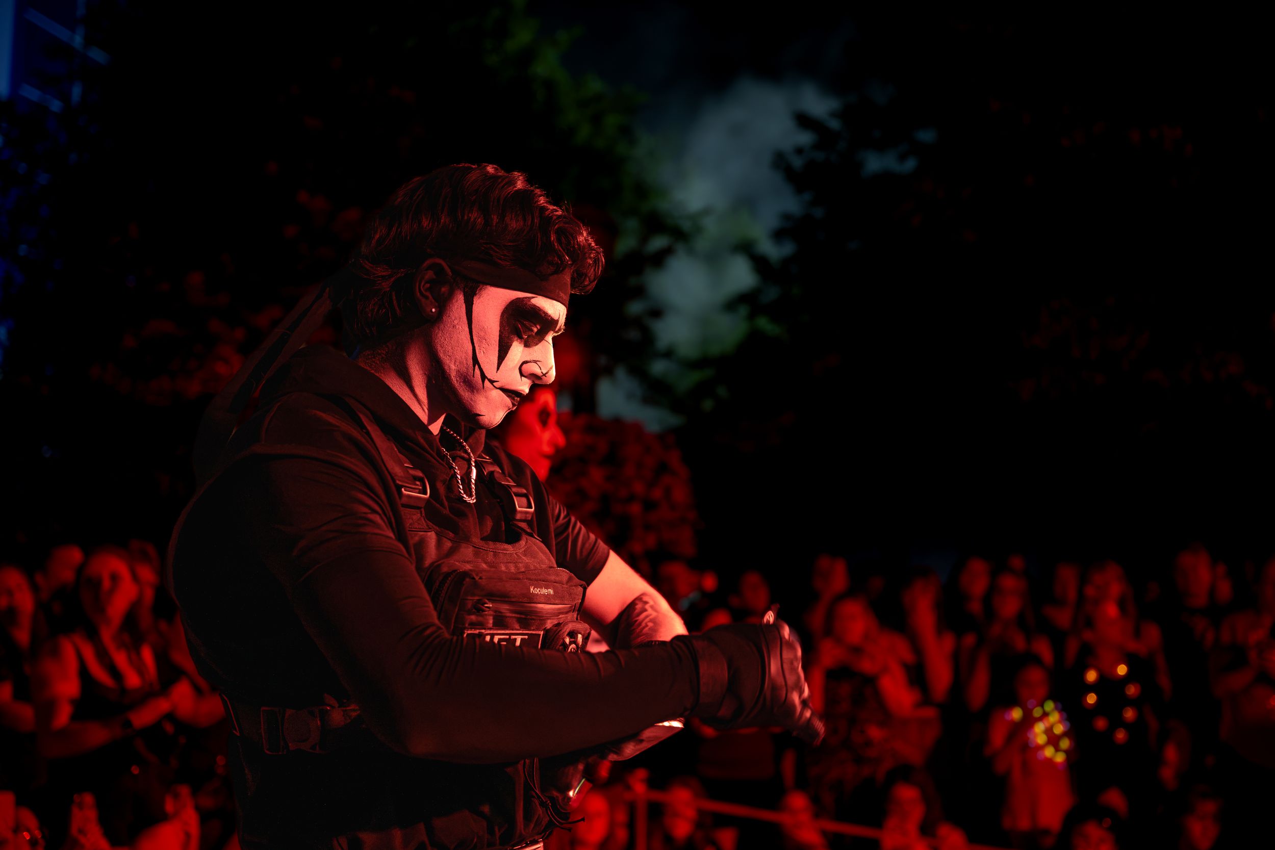 A performer in black costume and face paint plays guitar on stage before a large crowd at an outdoor concert.