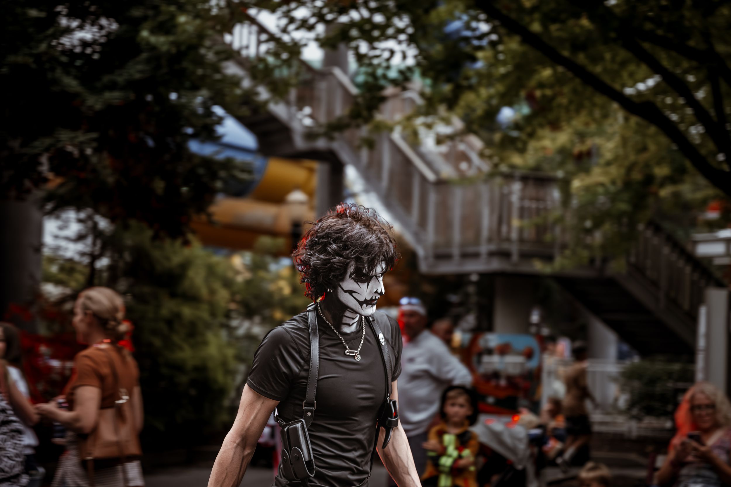 Person wearing a face mask walks through a crowded outdoor area with trees and a wooden structure in the background.