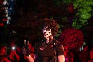 Person in skull face paint and black outfit waves at night Halloween event with red-lit trees in background.
