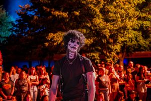 Person in creepy skull face paint costume stands before a crowd at an outdoor Halloween night event lit by orange lights.