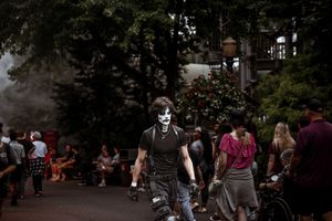 Person in skull face paint costume walks through a crowded Halloween theme park event on a foggy day.