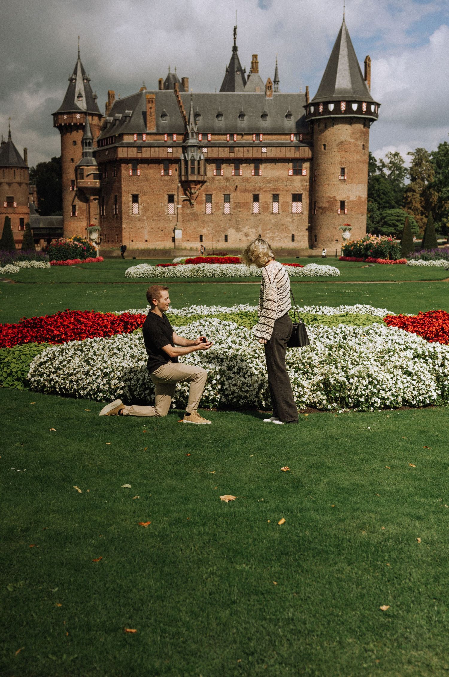 A man kneels on one knee proposing to a woman on a lawn with flower beds in front of a historic brick castle de Haar.