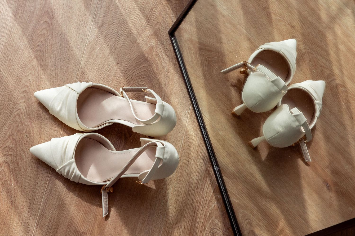Elegant cream pointed-toe heels with ankle straps on wood floor beside their mirror reflection.