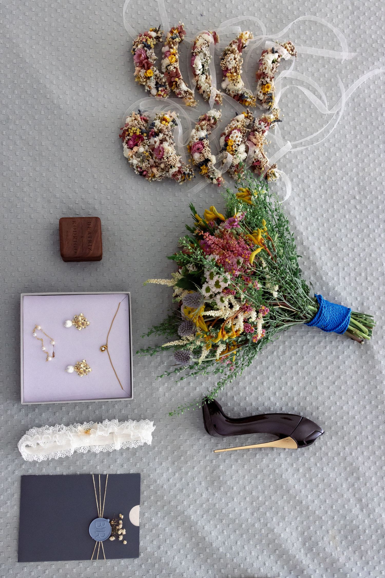 Flat lay of wedding accessories including a floral bouquet, high heel shoe, jewelry box, invitation, and dried flower letters.