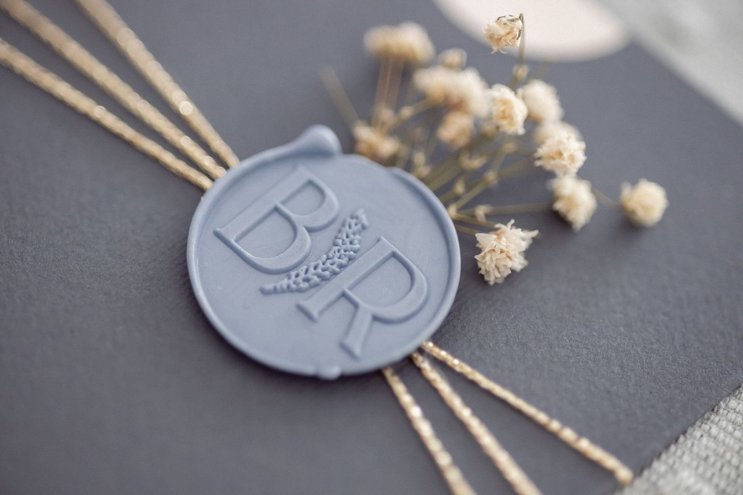 Elegant gray wax seal stamped with initials B and R, decorated with dried flowers on a dark gray surface.