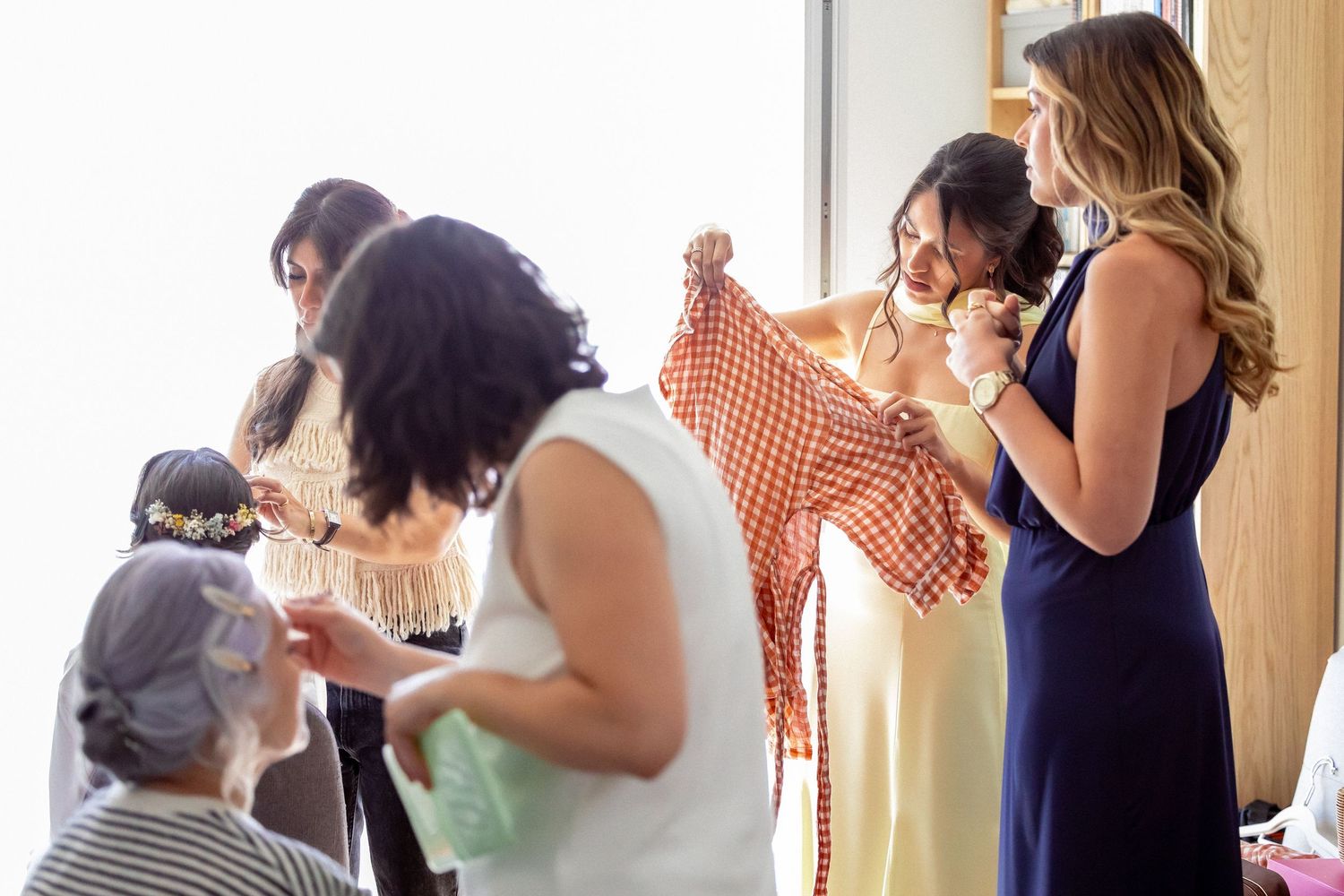 Bridesmaids helping bride prepare, examining red polka dot dress in bright room before wedding ceremony.