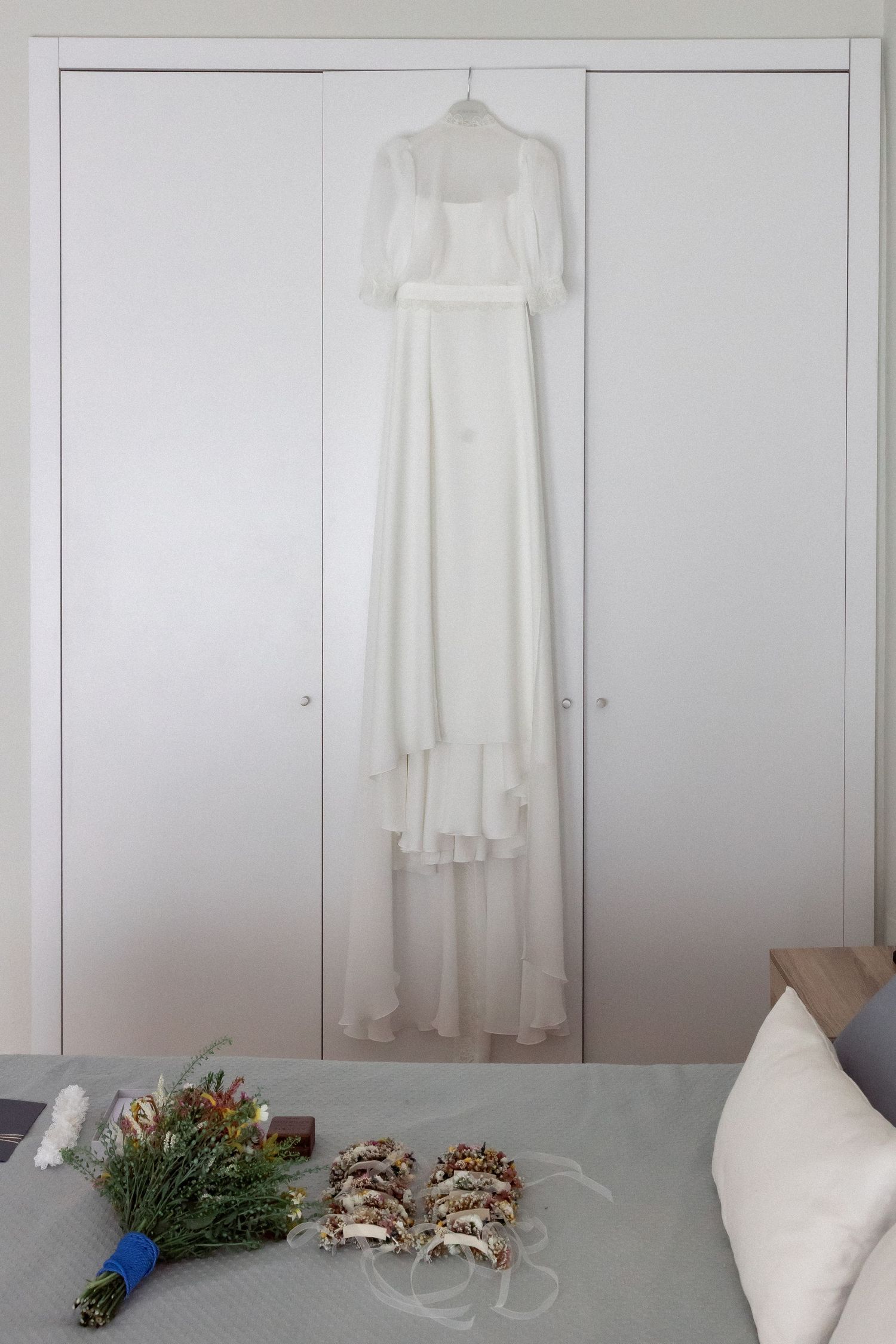 White wedding dress hanging on closet with bridal bouquet and sandals arranged on bed below.
