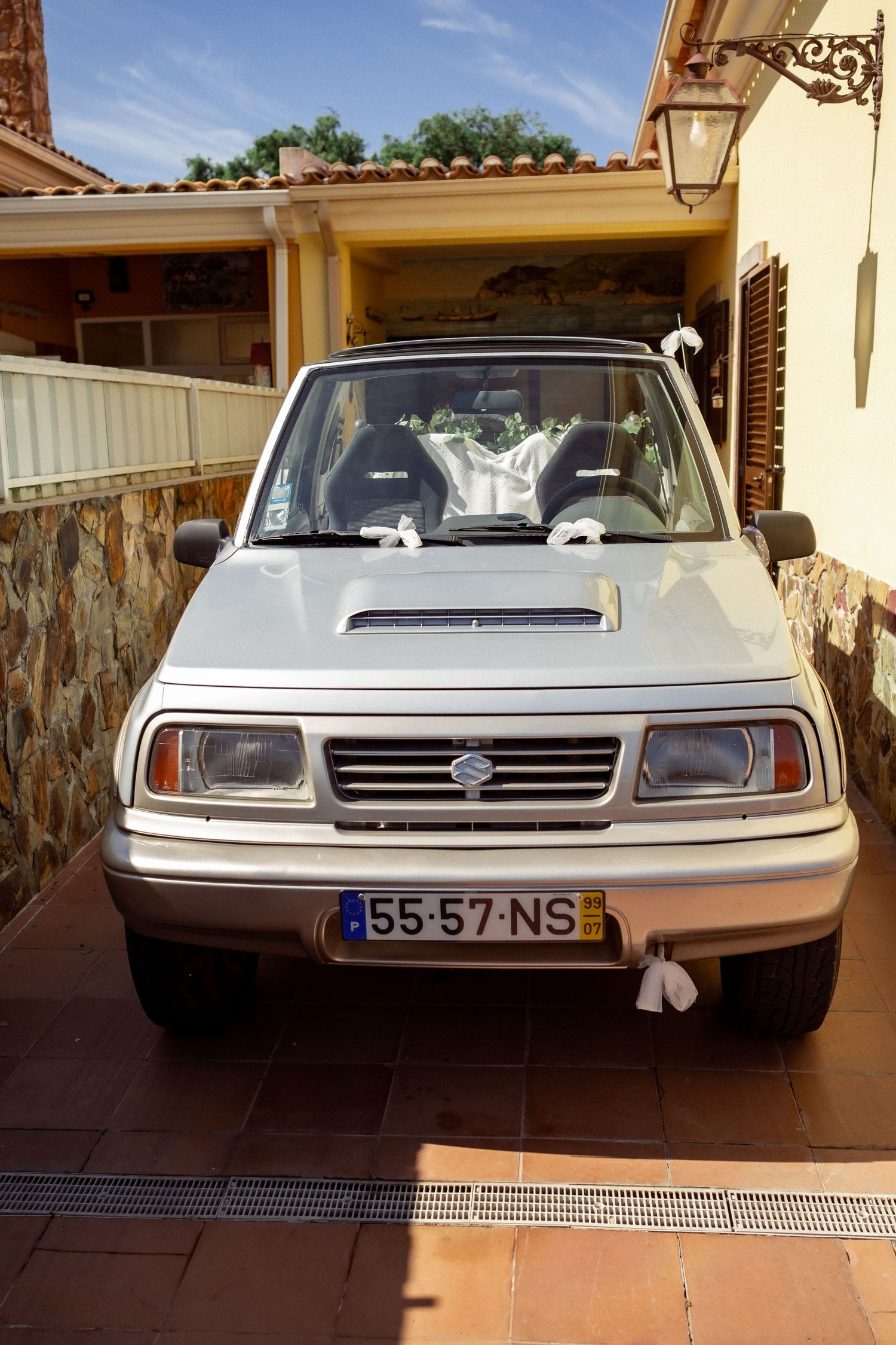 White Suzuki Vitara with license plate 55-57-NS decorated with wedding ribbons parked outside a stone building.