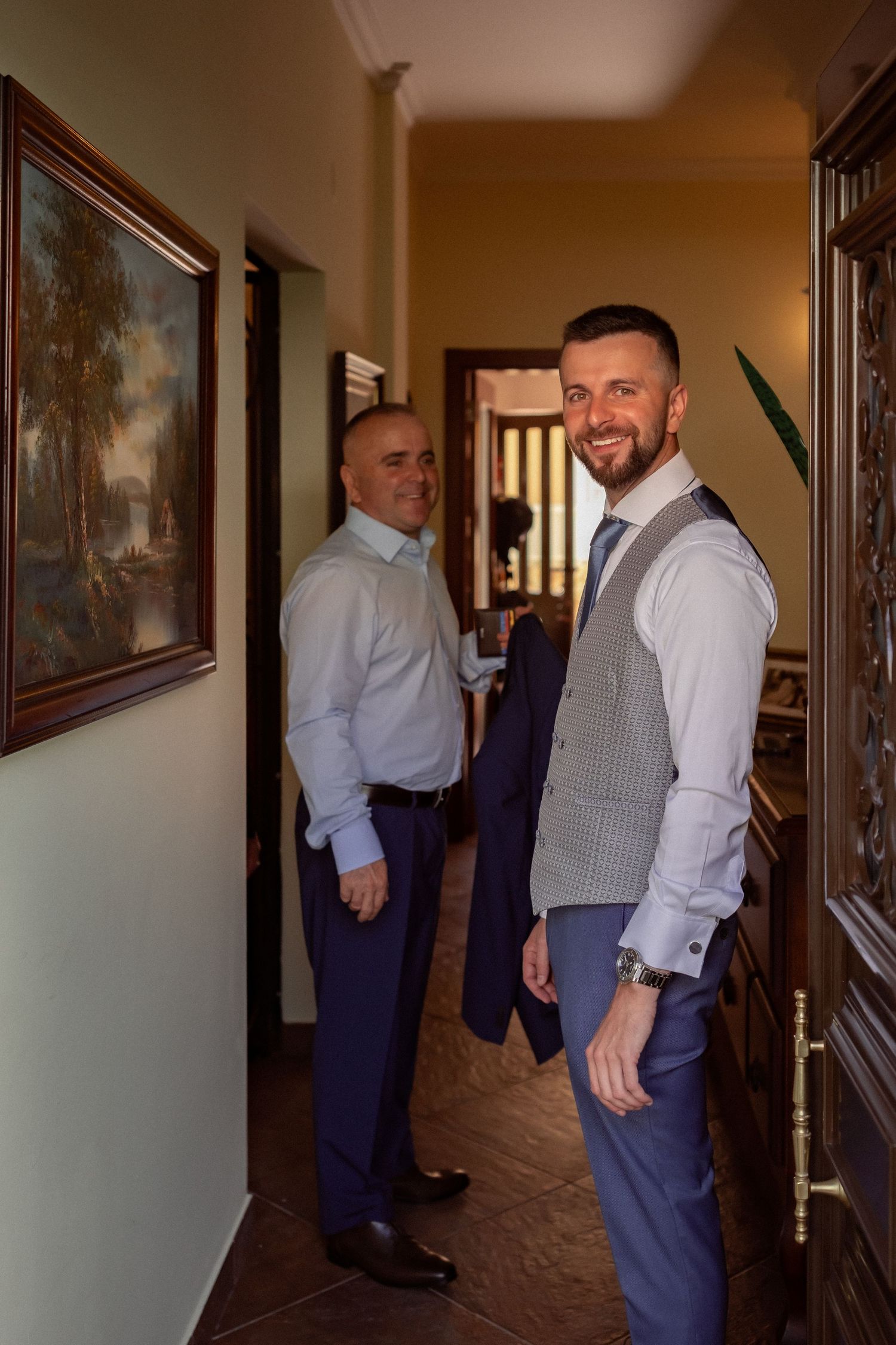 Two well-dressed men in suits stand in an elegant hallway with framed artwork, smiling at the camera.