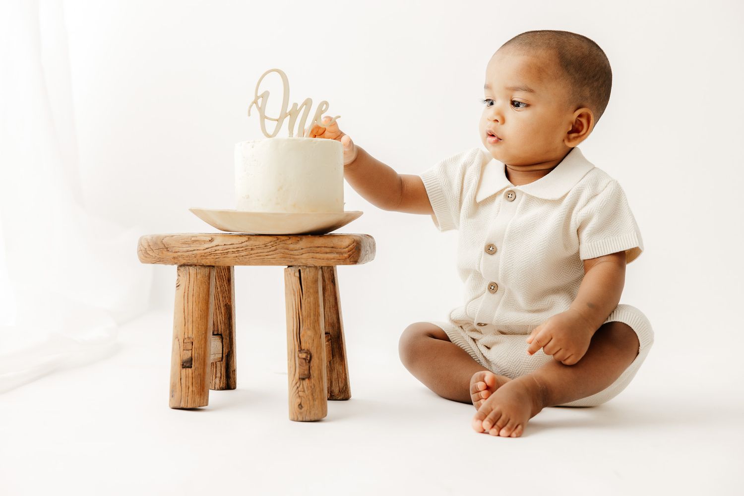 Baby in white outfit sits beside a large number 1 balloon for a first birthday photo on white background.