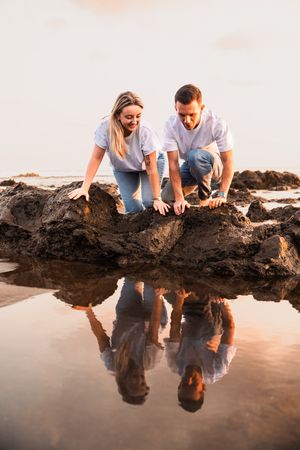Couple crouching on rocky beach at sunset, their reflections mirrored in a tidal pool below.
