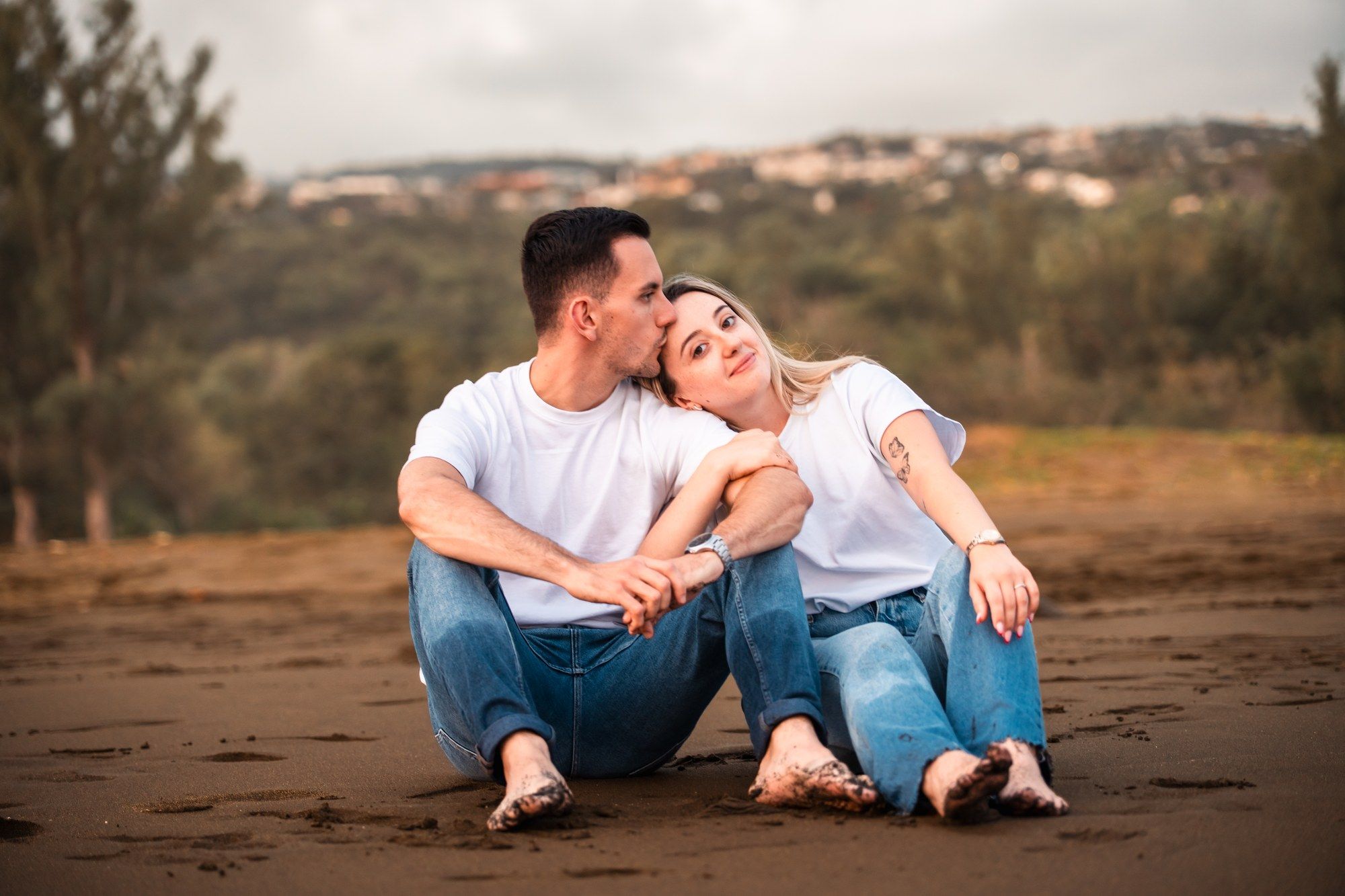 Romantic couple sitting on dark sand beach, woman leaning on mans shoulder, both wearing white tees and jeans.