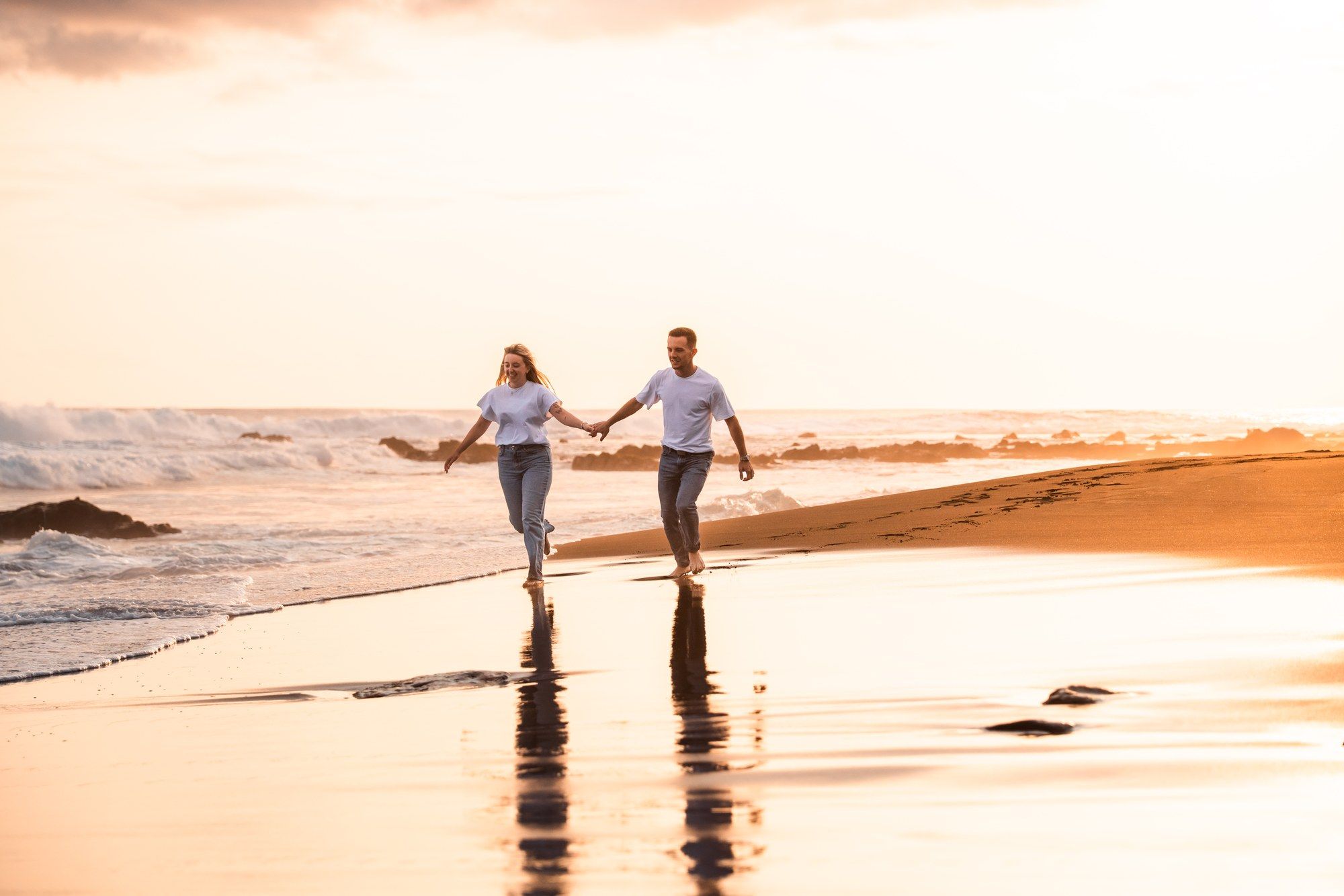 Couple holding hands running on a sandy beach at golden sunset with ocean waves in the background.