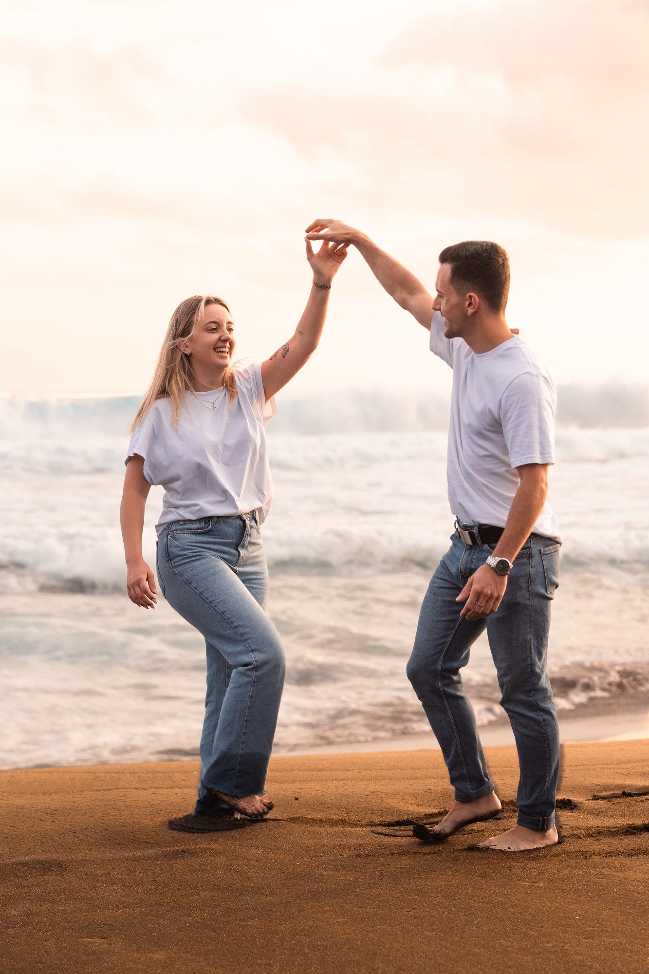 Couple dancing joyfully on a sandy beach at sunset, wearing matching white tees and jeans, holding hands near ocean waves.