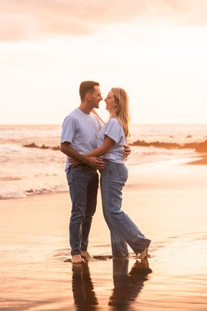 Romantic couple embracing barefoot on a beach at sunset, wearing matching white tees and jeans, with golden ocean waves behind them.