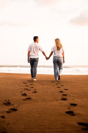 Couple holding hands walking on a sandy beach toward the ocean, leaving footprints in the sand.