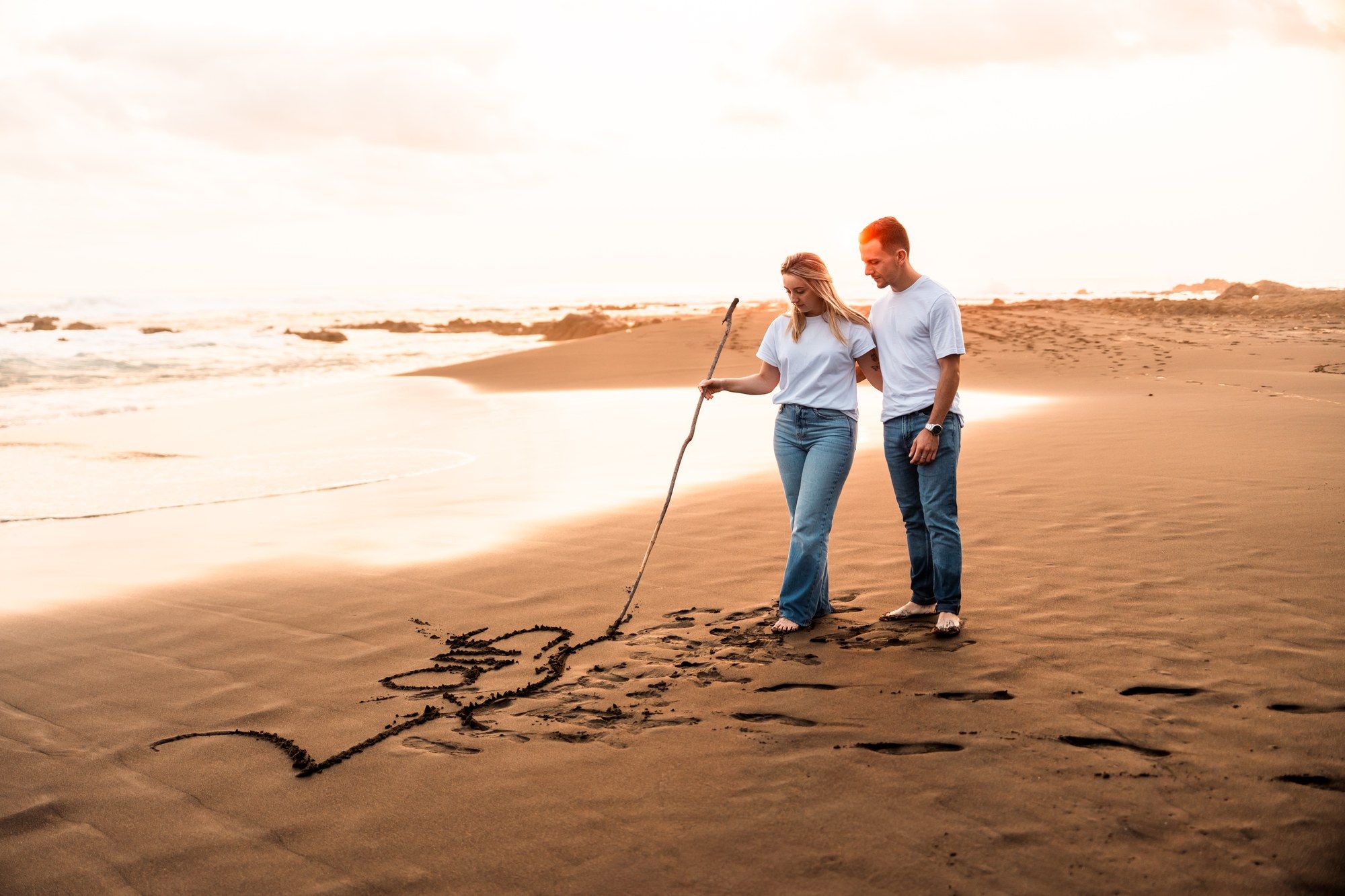Couple writing in sand on beach at sunset with stick, romantic golden hour seaside moment.