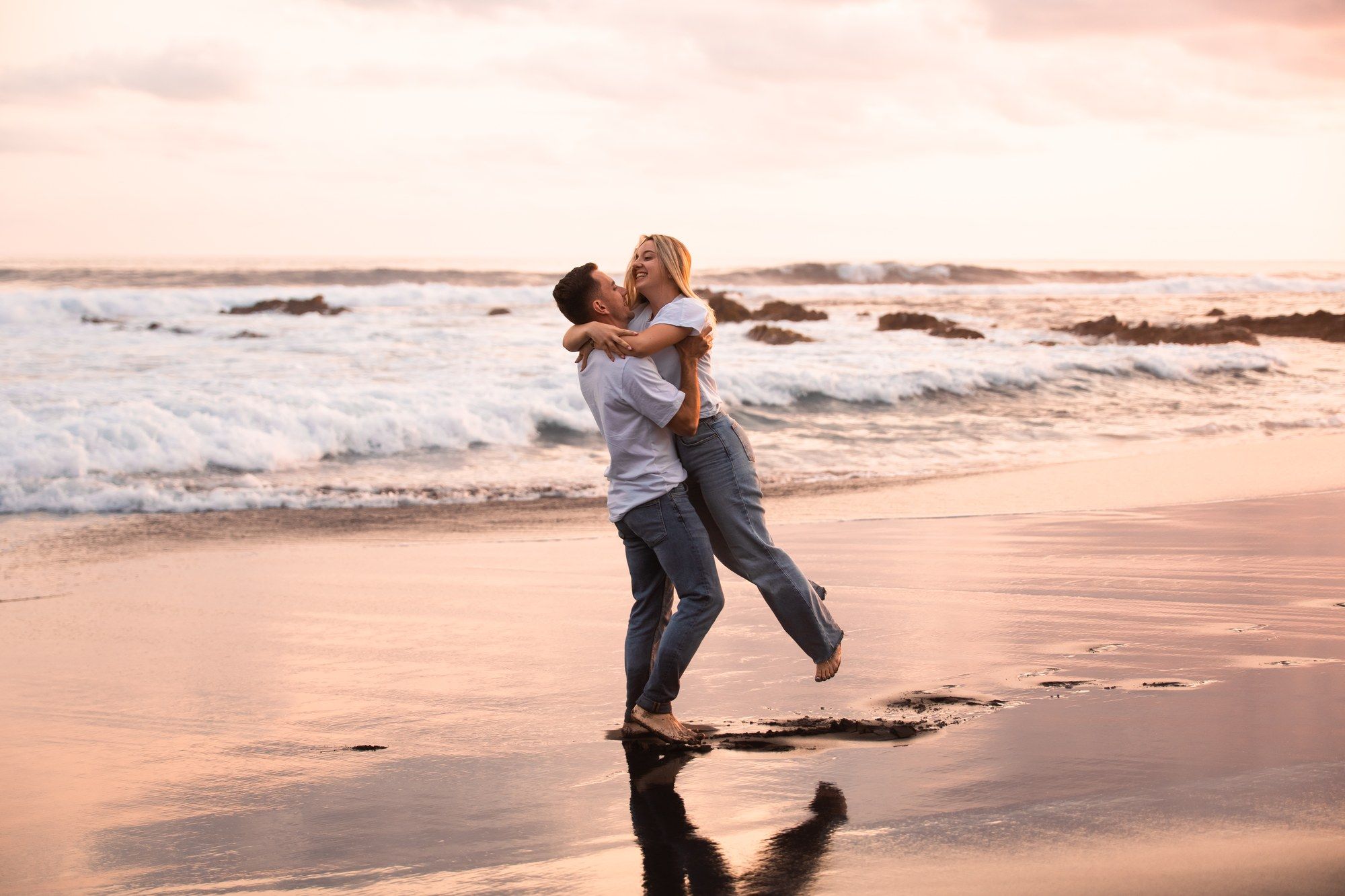 Couple embracing and spinning on a sandy beach at sunset with ocean waves in the background.