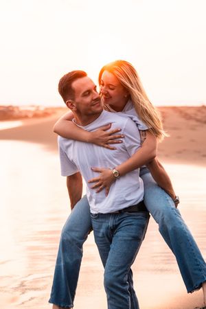 Couple sharing a romantic piggyback ride on a sandy beach at golden sunset, both wearing white shirts and jeans.