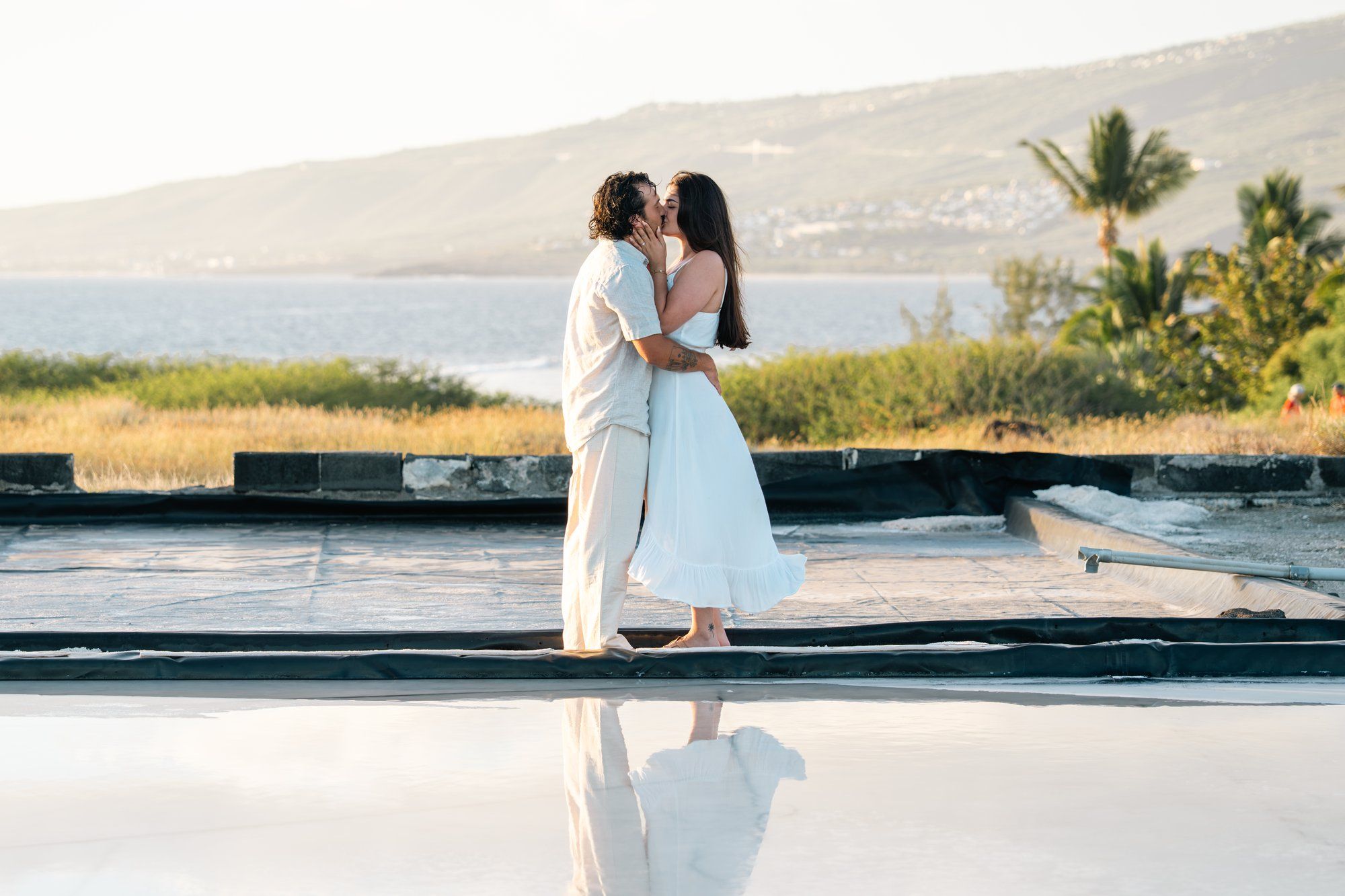 Couple sharing a romantic kiss in white attire by a reflective pool with ocean views at sunset.
