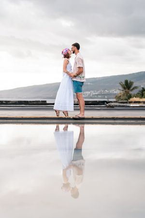 Couple kissing on a reflective surface near the ocean, woman in white dress with floral crown, man in blue shorts.
