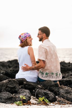 A couple sits on volcanic rocks by the ocean, smiling at each other, woman in colorful headscarf, man in tropical shirt.