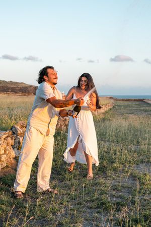 Couple in white outfits celebrating outdoors, popping champagne bottle in a sunny coastal field at golden hour.