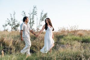 Couple holding hands walking through a grassy field in white outfits during golden hour.