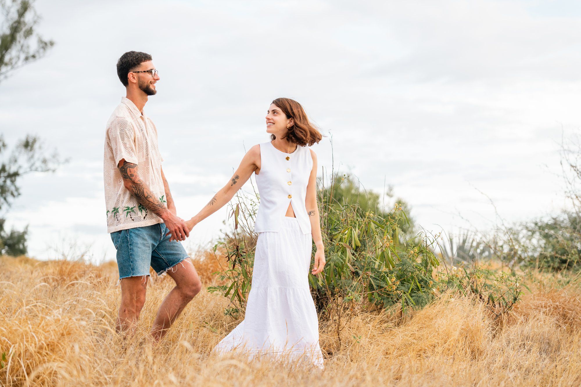 Couple holding hands in golden field, woman in white boho dress, man in casual attire, outdoor engagement photo.