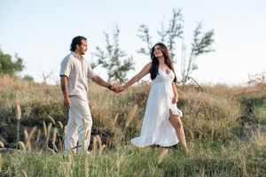 Couple holding hands walking through a grassy field, woman in white dress and man in white outfit, golden hour outdoor photo.