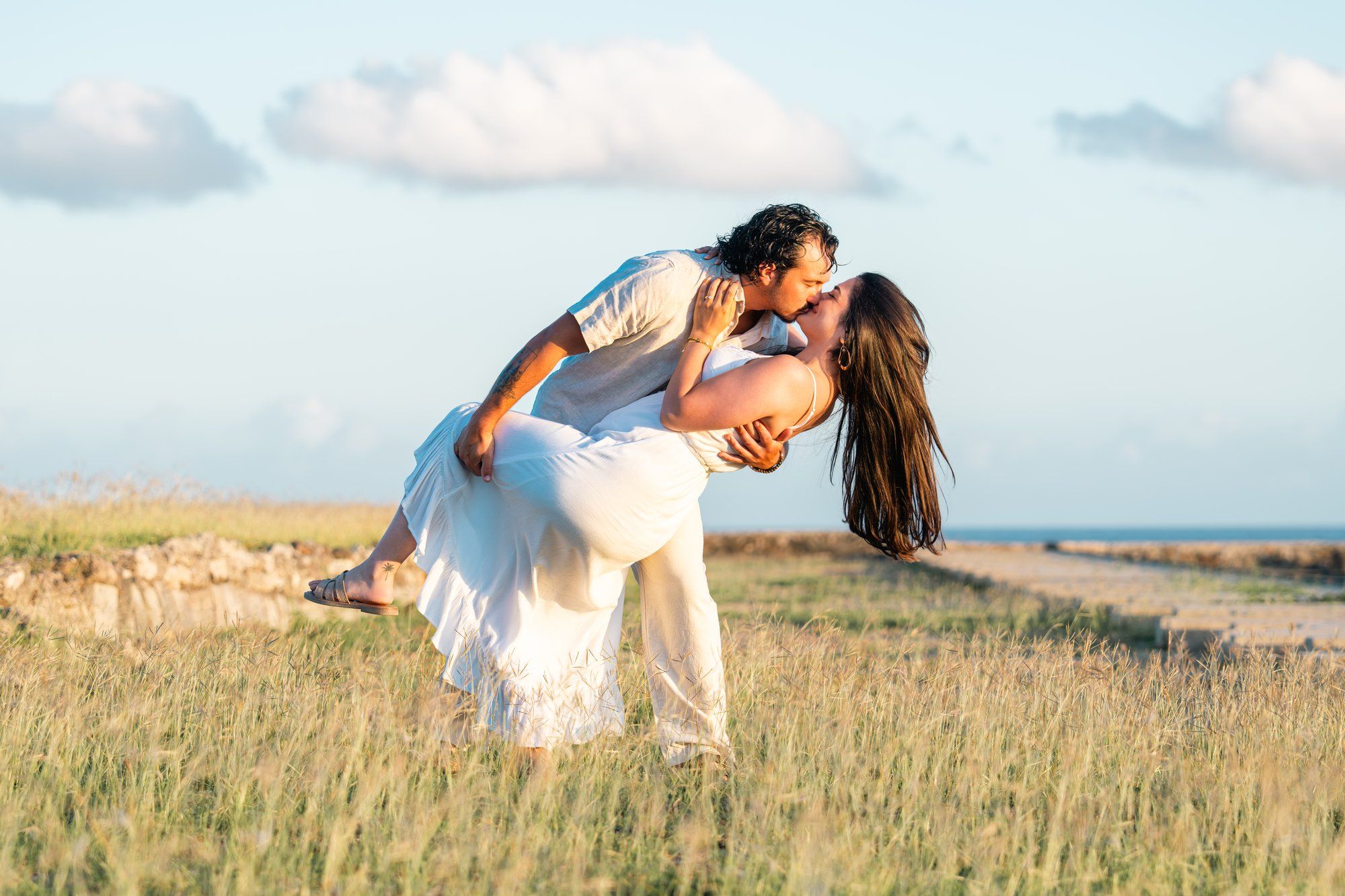 Couple sharing a romantic dip kiss in a sunny green field with a blue sky backdrop.