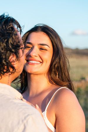 A smiling young woman with long brown hair enjoys a sunny outdoor moment with a partner in a golden field.