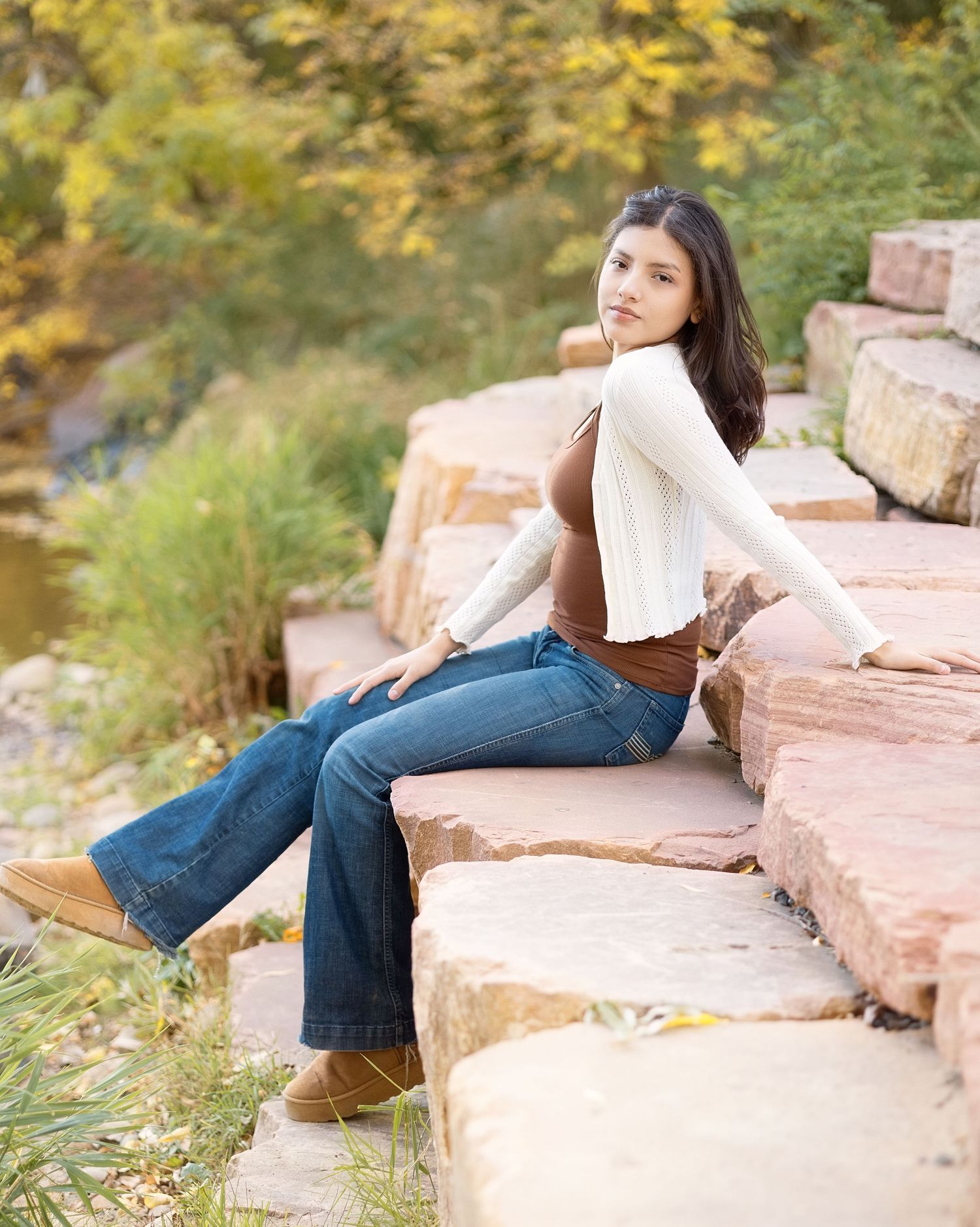 Young woman in flared jeans and white cardigan sitting on stone steps surrounded by autumn foliage.